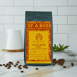 A bag of 'Calm Boss Lady' whole bean coffee with a yellow and black label, placed on a white surface with a ceramic cup and coffee beans scattered around.